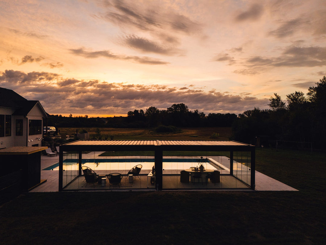 Anthracite-grey louvered pergola with glass walls lit from within at sunset, overlooking a residential pool and open countryside.