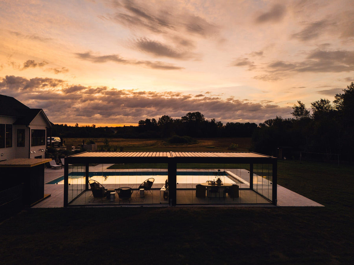 Anthracite-grey louvered pergola with glass walls lit from within at sunset, overlooking a residential pool and open countryside.